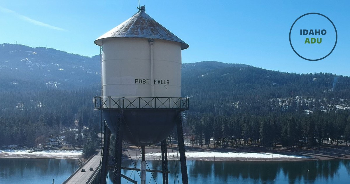 The historic Post Falls water tower, its tank lettered POST FALLS, standing above the Spokane River with forested hills behind it.